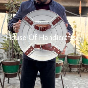 May include: A person holding a silver and brown Captain America shield. The shield has a star in the center and is made of metal. The shield is held in front of a person wearing a blue jacket.