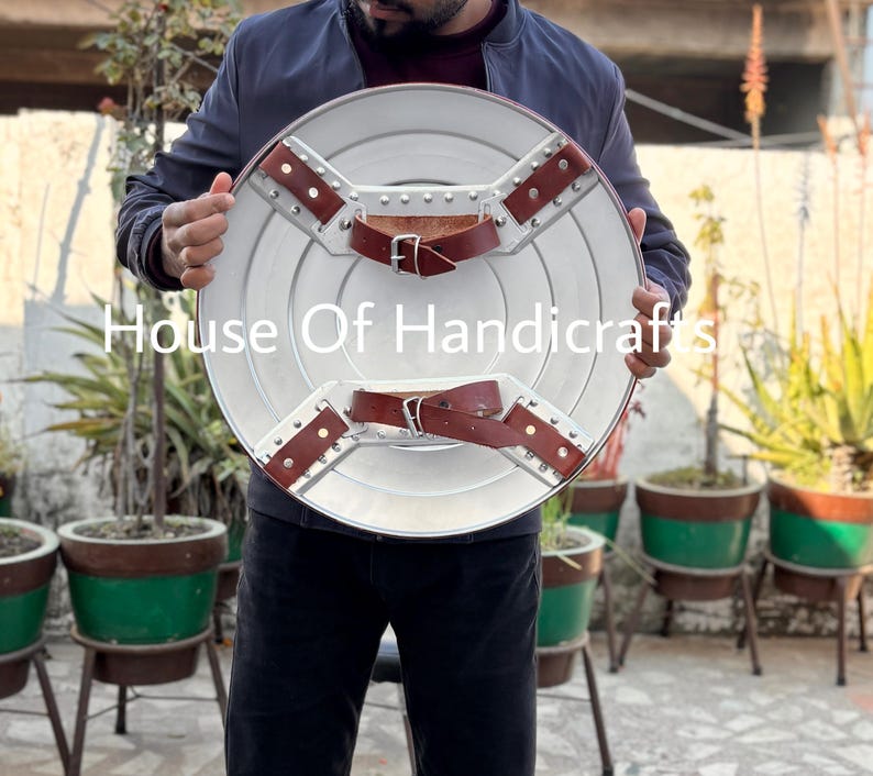 May include: A person holding a silver and brown Captain America shield. The shield has a star in the center and is made of metal. The shield is held in front of a person wearing a blue jacket.