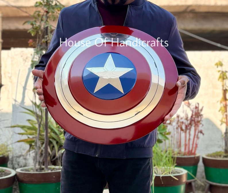 May include: A red, white, and blue Captain America shield with a silver metal center. The shield is being held by a person. The text "House Of Handicrafts" is visible in the image.