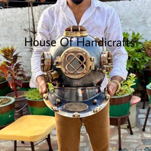 Può includere: Un casco da sub vintage in ottone e nero con un oblò in ottone e una valvola in ottone sul lato. Il casco è tenuto da una persona che indossa una camicia bianca e pantaloni marroni. Il testo "House Of Handicrafts" è visibile nell'immagine.