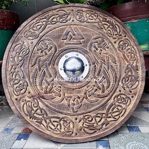 以下が含まれることがあります： 複雑な結び目模様の彫刻と中央の金属製ボスが特徴の円形木製シールド。中央にはHouse Of Handicraftsの文字が見える。木材はダークブラウン仕上げで、表面全体に詳細な彫刻が施されている。