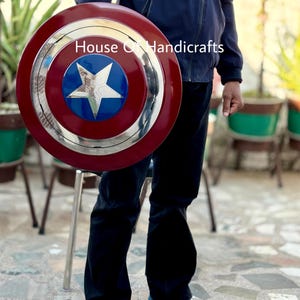 May include: A red, white, and blue Captain America shield with a silver star in the center. The shield is made of metal and has a raised rim. The shield is being held by a person wearing a blue jacket and jeans. The text "House of Handicrafts" is visible in the image.