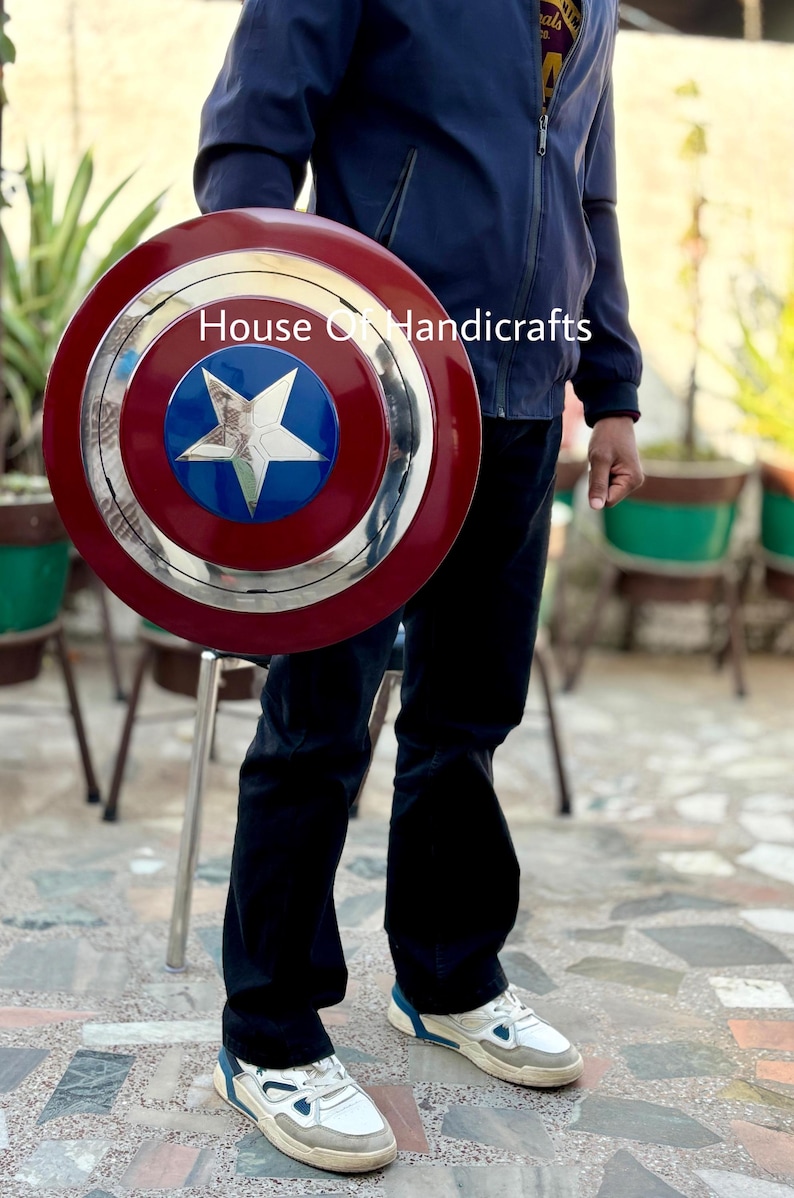 May include: A red, white, and blue Captain America shield with a silver star in the center. The shield is made of metal and has a raised rim. The shield is being held by a person wearing a blue jacket and jeans. The text "House of Handicrafts" is visible in the image.