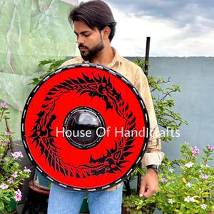 Könnte beinhalten: Ein runder, roter Schild mit einem schwarzen Drachendesign, das die Mitte umkreist. Der Schild hat einen schwarzen Rand und der Text "House Of Handicrafts" ist auf der Vorderseite aufgedruckt. Der Schild wird von einer Person gehalten.