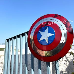 May include: A red, white, and blue Captain America shield with a silver metal rim. The shield has a five-pointed star in the center. The shield is hanging on a blue metal fence. The text "House Of Handicrafts" is visible in the upper right corner of the image.