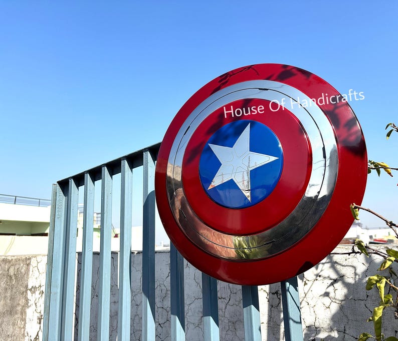 May include: A red, white, and blue Captain America shield with a silver metal rim. The shield has a five-pointed star in the center. The shield is hanging on a blue metal fence. The text "House Of Handicrafts" is visible in the upper right corner of the image.