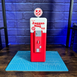 May include: A red and white Jugger-Nog vending machine replica featuring a circular top sign with a white cross icon. The front displays the text Drink Jugger-Nog 10 cents Made with real eggs, with a grey central panel containing small rectangular details.