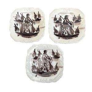 May include: Three square ceramic plates with scalloped edges, each featuring a hand-painted dark brown illustration of a figure carrying a long tool through tall grass. The plates have a cream-colored base with embossed decorative borders.
