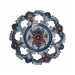 May include: A decorative ceramic plate with a scalloped edge and intricate floral designs. The plate features a central floral motif with red, yellow, and green accents, surrounded by a blue and white border. Additional red flowers are placed around the edge.