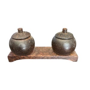 May include: Two dark brown, spherical containers with matching lids, each featuring a textured, speckled surface. These round vessels are positioned side-by-side on a rectangular, flat wooden base that has a visible grain and a smooth finish.