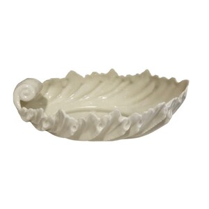 May include: A glossy off-white ceramic dish shaped like a curled leaf. The bowl features a smooth interior surface and a textured, scalloped edge that mimics the appearance of foliage, with a prominent spiral detail on one side of the rim.