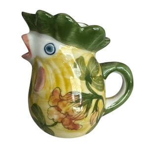 May include: A ceramic creamer shaped like a rooster, featuring a green comb, a yellow body with painted orange and yellow floral patterns, a blue eye, and a curved handle. The pitcher has an open beak spout and a smooth, glossy finish.