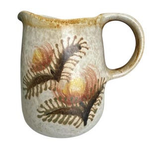 May include: A ceramic pitcher with a speckled beige and cream glaze, featuring a hand-painted floral design in shades of yellow, orange, and dark brown. The pitcher has a curved handle, a wide spout, and a textured finish across the entire surface.