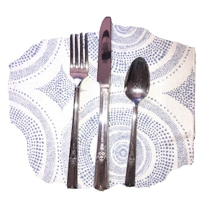 May include: A stainless steel fork, knife, and spoon set on a white and blue patterned napkin. The silverware has decorative detailing on the handles. The napkin has a circular design with small blue dots.