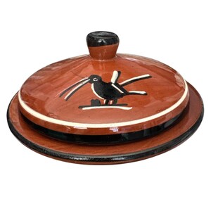 May include: A ceramic butter dish with a lid. The dish is a reddish-brown color with a black and white bird design on the lid. The rim of the dish has a white stripe, and the base has a black stripe. The lid has a black knob.