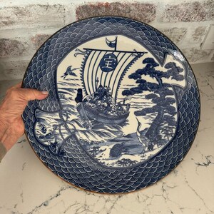 May include: A round ceramic plate featuring a blue and white illustration of a traditional sailing ship with the kanji character 宝 on its sail. The design includes a pine tree, a crane, a turtle, and a border pattern of stylized ocean waves.