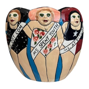 May include: A ceramic vase featuring three figures in swimsuits and sunglasses. Each figure has a sash with text: "MS. DOT COM", "MS. SPENT YOUTH", and "MS. ATTITUDE". The vase has a rounded shape with blue and black accents.