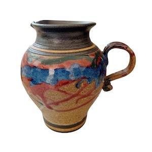 May include: A ceramic pitcher with a rounded body and a single handle. The surface features a textured tan base with horizontal bands of blue and reddish-brown glaze. The neck and rim are finished in a dark, solid black glaze with a smooth texture.