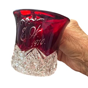 May include: A ruby red glass tumbler featuring a clear, diamond-cut base and an etched design on the upper portion. The engraving includes decorative flourishes above the initials E.H. and the year 1910, all rendered in a white, frosted style.