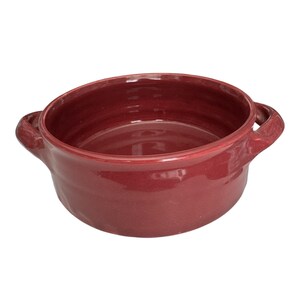 May include: A deep, round ceramic bowl with a glossy, dark red finish. The bowl features two small, integrated handles on opposite sides, a smooth interior surface, and a wide, open top, presenting a solid, uniform color throughout the exterior.