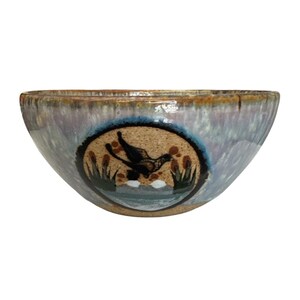 May include: A ceramic bowl with a mottled lavender and blue glaze featuring a central circular medallion. The medallion depicts a black bird in flight over a marshy landscape with green reeds and white accents against a speckled tan background.