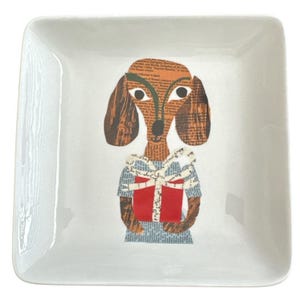 May include: A square white ceramic dish featuring a collage-style illustration of a brown dog with floppy ears. The dog is depicted holding a bright red gift box tied with a white ribbon, with the body composed of printed newspaper text.