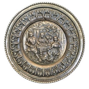 May include: A round metallic decorative plate featuring a central embossed scene of four figures gathered around a table. The border is decorated with a repeating floral pattern and a twisted rope edge. The finish is a muted, aged silver tone.