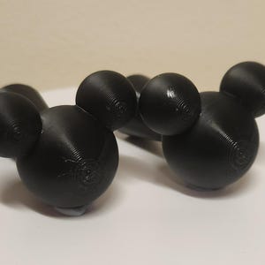 May include: Two black 3D printed objects, each featuring a large central sphere with two smaller spheres attached to the top, resembling a mouse head silhouette. The surfaces show fine horizontal layer lines from the additive manufacturing process.