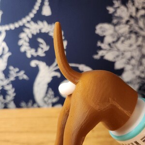 May include: A brown 3D printed dog figurine with a white tail. The dog is holding a tube of toothpaste in its back legs.