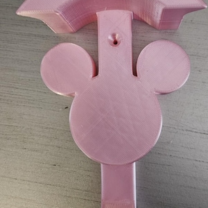 May include: A pink plastic wall hook shaped like Mickey Mouse's head. The hook has a rounded top and a single hook at the bottom.