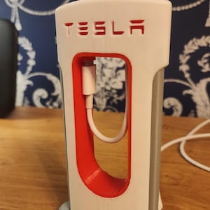 May include: A white 3D printed Tesla charging station stand with a red charging port and the word "TESLA" printed in red on the front. A white charging cable is threaded through the charging port.