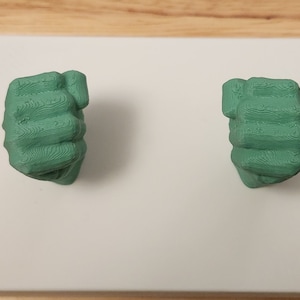 May include: Two green 3D printed fists, clenched and facing forward, are mounted on a white surface.
