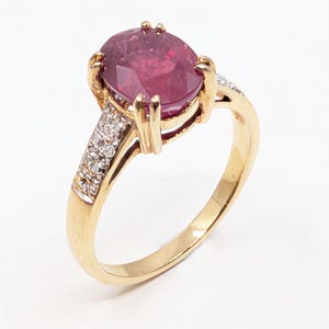 May include: A gold ring featuring a large, oval-cut ruby gemstone. The ruby is a deep red color and is the focal point of the ring. The band is gold, with small accent diamonds set on either side of the ruby.