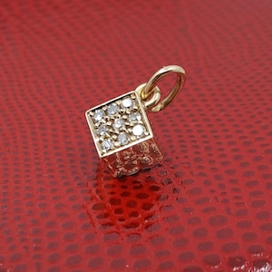 Könnte beinhalten: Ein goldener Anhänger in Form eines Würfels mit kleinen Diamanten, die in die Oberfläche eingefasst sind. Der Anhänger hat eine Öse oben zum Anbringen an einer Halskette oder einem Armband.