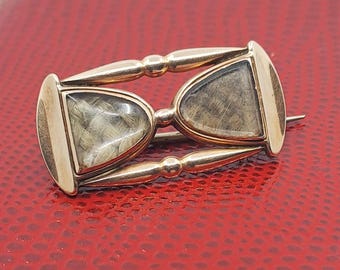May include: A gold-toned hourglass brooch with two glass-covered compartments filled with a light-colored material. The brooch has a pin on the back and is set against a red background.