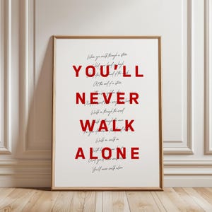 Puede incluir: Una lámina enmarcada con la letra de la canción You'll Never Walk Alone. El texto se presenta en dos estilos: letras mayúsculas rojas, grandes y en negrita que forman YOU'LL NEVER WALK ALONE, superpuestas sobre una letra cursiva negra más pequeña.