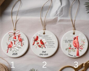 May include: Three round ceramic Christmas ornaments with gold string hangers. Each ornament features a red cardinal bird design with text. One says "2025", another says "Dad 1943-2025", and the last says "I AM ALWAYS WITH YOU Michelle-2025".