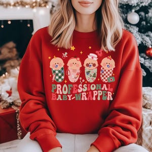 Puede incluir: Sudadera roja con el texto "PROFESSIONAL BABY-WRAPPER" en rosa y verde. El gráfico presenta cuatro bebés de dibujos animados envueltos en mantas estampadas, cada uno con un chupete. El diseño incluye estrellas e iconos de bebé.
