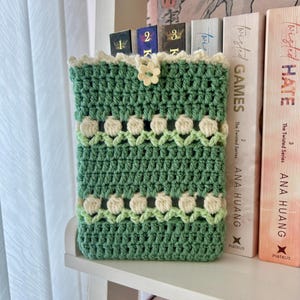 May include: A green crochet book sleeve with a white floral pattern and a button fastening. The sleeve, made from textured yarn, protects books. It's displayed on a white shelf beside books. The sleeve is approximately 20 cm tall.