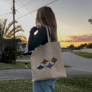 以下が含まれることがあります： 人が、ベージュのトートバッグを肩にかけています。バッグには、茶色、青、グレーの3匹のスタイリッシュなエイがデザインされています。バッグには長いストラップが付いています。