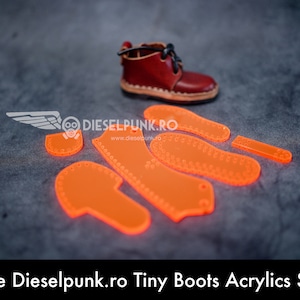 Peut inclure: Un ensemble de gabarits en acrylique orange pour la fabrication de petites bottes. Les gabarits sont disposés sur une surface grise à côté d'une paire de mini-bottes en cuir marron. Le texte "Dieselpunk.ro Tiny Boots Acrylics Set" est visible en bas de l'image.