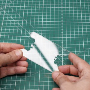 May include: A clear plastic stencil with a jagged edge design. The stencil is being held by two hands.