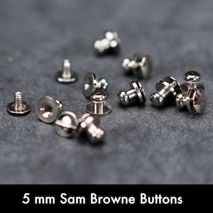 Peut inclure: Gros plan sur un groupe de boutons Sam Browne argentés de 5 mm. Les boutons sont éparpillés sur une surface gris foncé.