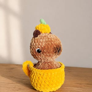 May include: A brown crocheted capybara plush toy with a small yellow pumpkin and green stem on its head, sitting inside a bright yellow crocheted mug with a handle. The plush has a textured, soft yarn appearance with black plastic eyes.