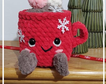 May include: A red crocheted mug plushie featuring a smiling face with black eyes, pink cheeks, and brown dangling legs. The mug is topped with pink and white crocheted pom-poms and decorated with white snowflake accents on the exterior surface.