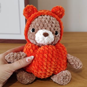 May include: A plush stuffed animal crafted from soft, textured yarn. The figure features a brown bear head with a white muzzle and a dark brown nose, wearing a bright orange hooded outfit with small rounded ears attached to the hood.