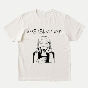 Pode incluir: Uma t-shirt de manga curta na cor creme com uma estampa gráfica preta de um personagem sorridente e barbudo segurando uma chávena. Acima do personagem, o texto "MAKE TEA, NOT WAR." está escrito numa fonte escura de estilo manual.