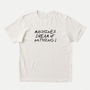 May include: A short-sleeved, off-white crew neck t-shirt featuring the black text "MACHINES DREAM OF NOTHING!" printed in a handwritten style across the centre of the chest. The garment has a standard fit with a ribbed collar and hemmed sleeves.