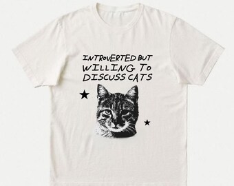 Puede incluir: Una camiseta de manga corta color blanco roto con un gráfico negro de una cara de gato centrado en el pecho. Sobre el gato, el texto dice INTROVERTED BUT WILLING TO DISCUSS CATS en una fuente negra, flanqueado por dos pequeñas estrellas.