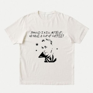 Könnte beinhalten: Ein cremefarbenes Kurzarm-T-Shirt mit einem schwarzen Grafikdruck eines Gesichts, drei kleinen Sternen und dem Text "SHOULD I KILL MYSELF, OR HAVE A CUP OF COFFEE?" in einer fetten, handgezeichneten Schriftart auf der Brustpartie.